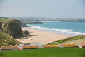 A coastal landscape with a beach, cliffs, and a view of a distant city. - 4 Bed in Watergate Bay (oc-b584373) (Tregurrian)