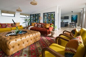 A modern living room with a large leather ottoman, yellow and red sofas, and a dining area with a table and chairs. - 4 Bed in Watergate Bay (oc-b584373) (Tregurrian)