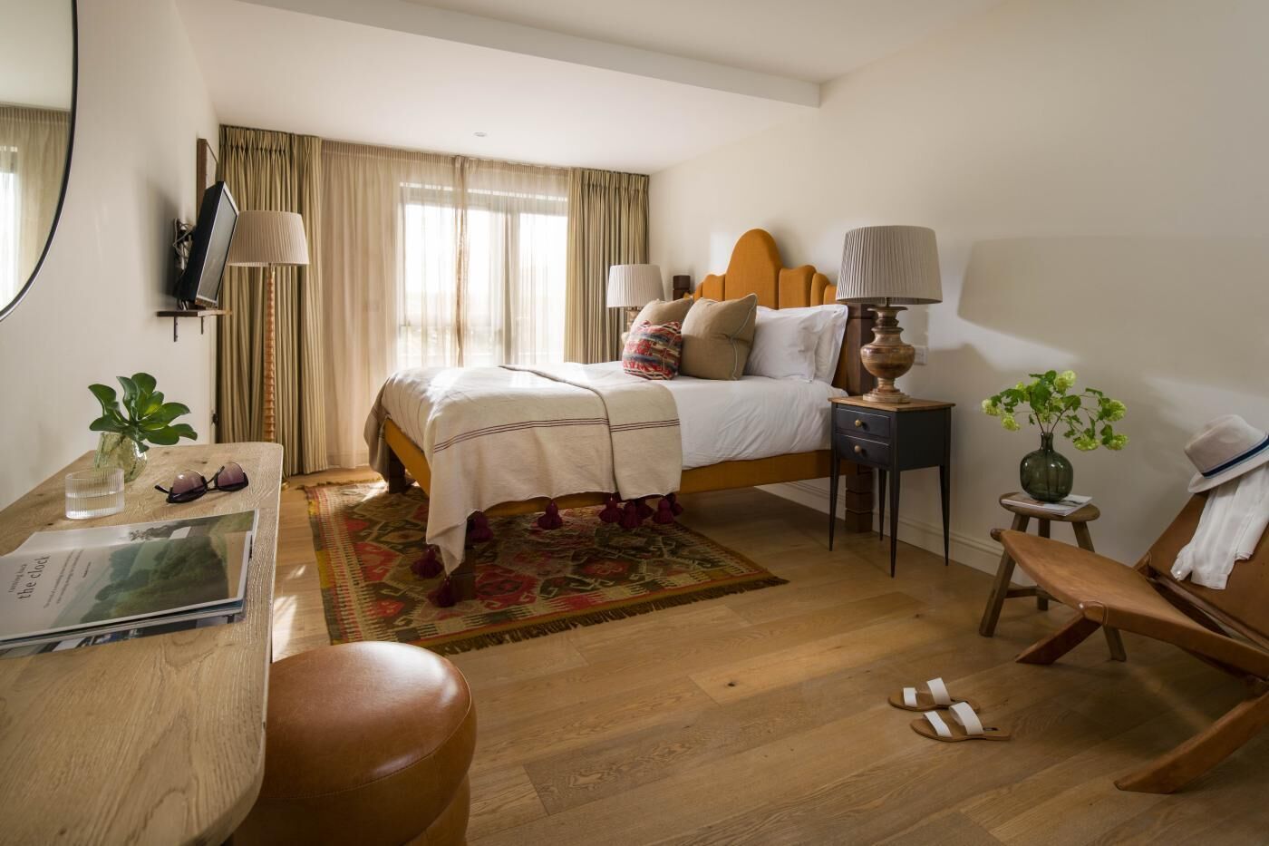 Ein Hotelzimmer mit einem Bett, Nachttischen, einem Schreibtisch, einem Sessel, einem kleinen Tisch mit einer Vase und einem Vorhangfenster.