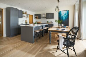 A modern kitchen with a central island, bar stools, and a dining table with chairs. - 4 Bed in Watergate Bay (oc-b584373) (Tregurrian)