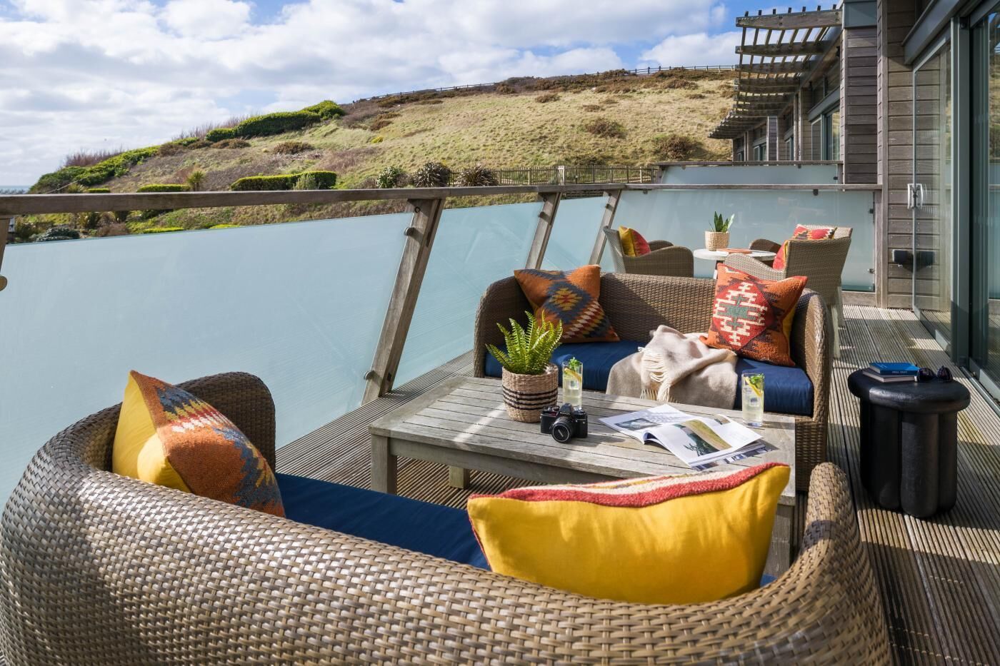 Une terrasse dotée de meubles en osier, d’une table avec des magazines et offrant une vue sur un lac et des collines.