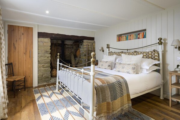 Une chambre avec un mur en pierre, un lit blanc agrémenté d’oreillers à motifs, une chaise en bois et un tapis.