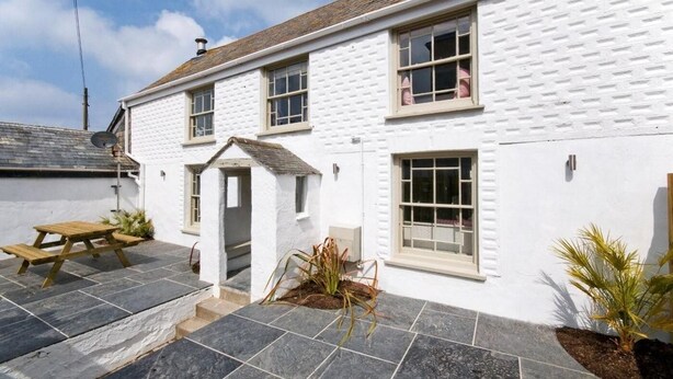 A white building with a tiled patio, a wooden bench, and a small garden area. - 5 Bed in Porth (oc-b584392) (Newquay)