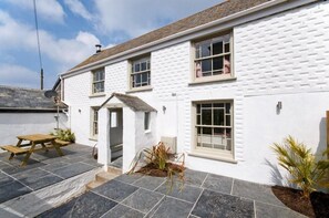 A white building with a tiled patio, a wooden bench, and a small garden area. - 5 Bed in Porth (oc-b584392) (Newquay)