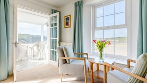 A room with a view of the sea, a white chair, a wooden chair, a small table with a vase of flowers, and a door leading outside. - 2 Bed in Porth (oc-b584410) (Newquay)