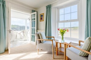 Ein Zimmer mit Meerblick, ein weißer Stuhl, ein hölzerner Stuhl, ein kleiner Tisch mit Blumenvase und eine Tür, die nach draußen führt.