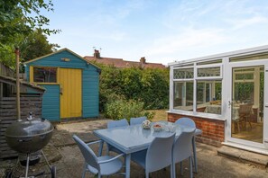 Une terrasse avec une table et des chaises, un barbecue, un abri de jardin et une pièce fermée par des baies vitrées.