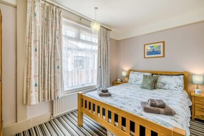 A bedroom with a bed, a window with curtains, a radiator, a bedside table with a lamp, and a framed picture on the wall. - 3 Bed in Southwold (oc-hseami) (Southwold)