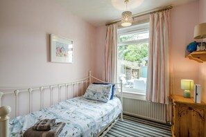 Une chambre à coucher avec un lit, une fenêtre avec des rideaux, une lampe de chevet, un radiateur et une petite armoire en bois.