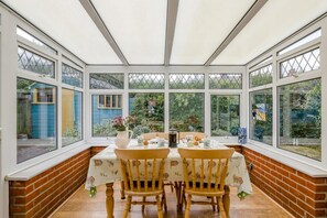 Ein Wintergarten mit Backsteinwand, zwei Holzstühlen, einem Tisch mit weißem Tischleinen und Blick auf einen Garten.