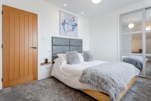 Ein Schlafzimmer mit Holztür, grau gepolstertem Kopfteil, einem weißen Bett mit grauen Kissen und einer flauschigen grauen Decke. An der Wand hängt ein abstraktes Gemälde.