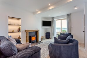 Ein modernes Wohnzimmer mit Kamin, bequemen Sitzmöglichkeiten, einem Flachbildfernseher und Blick ins Freie.