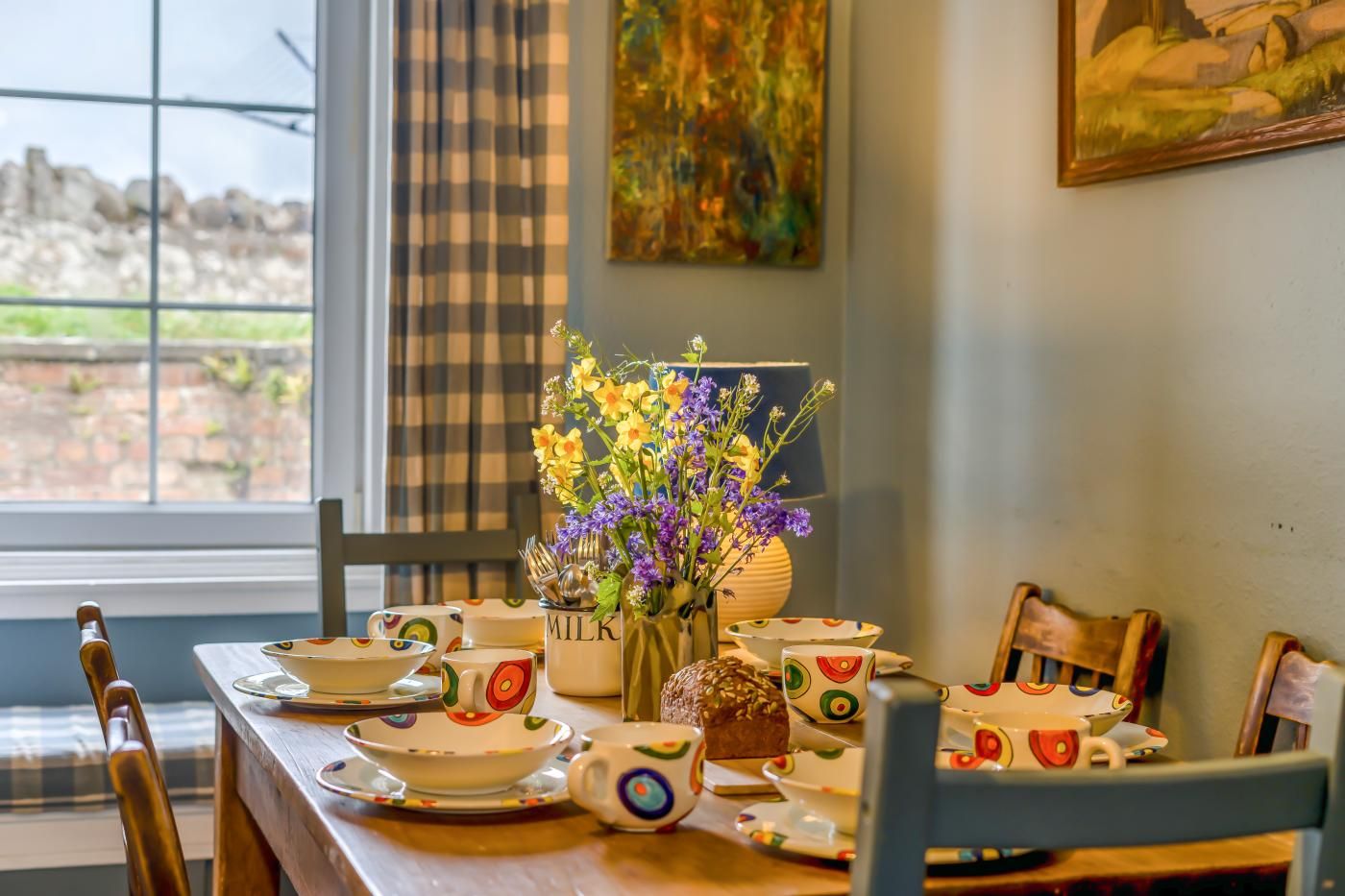 Mesa de jantar com louça colorida e um arranjo floral, em um ambiente com janela e obras de arte na parede.