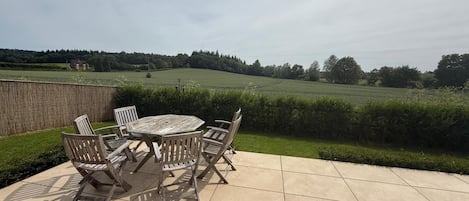 Un patio exterior con una mesa redonda y seis sillas, con vista a un campo de césped y una casa distante.