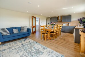A modern living room with a blue sofa, a dining table with chairs, and a kitchen area with cabinets and a stove. - 4 Bed in Longforgan (oc-w42661) (Longforgan)