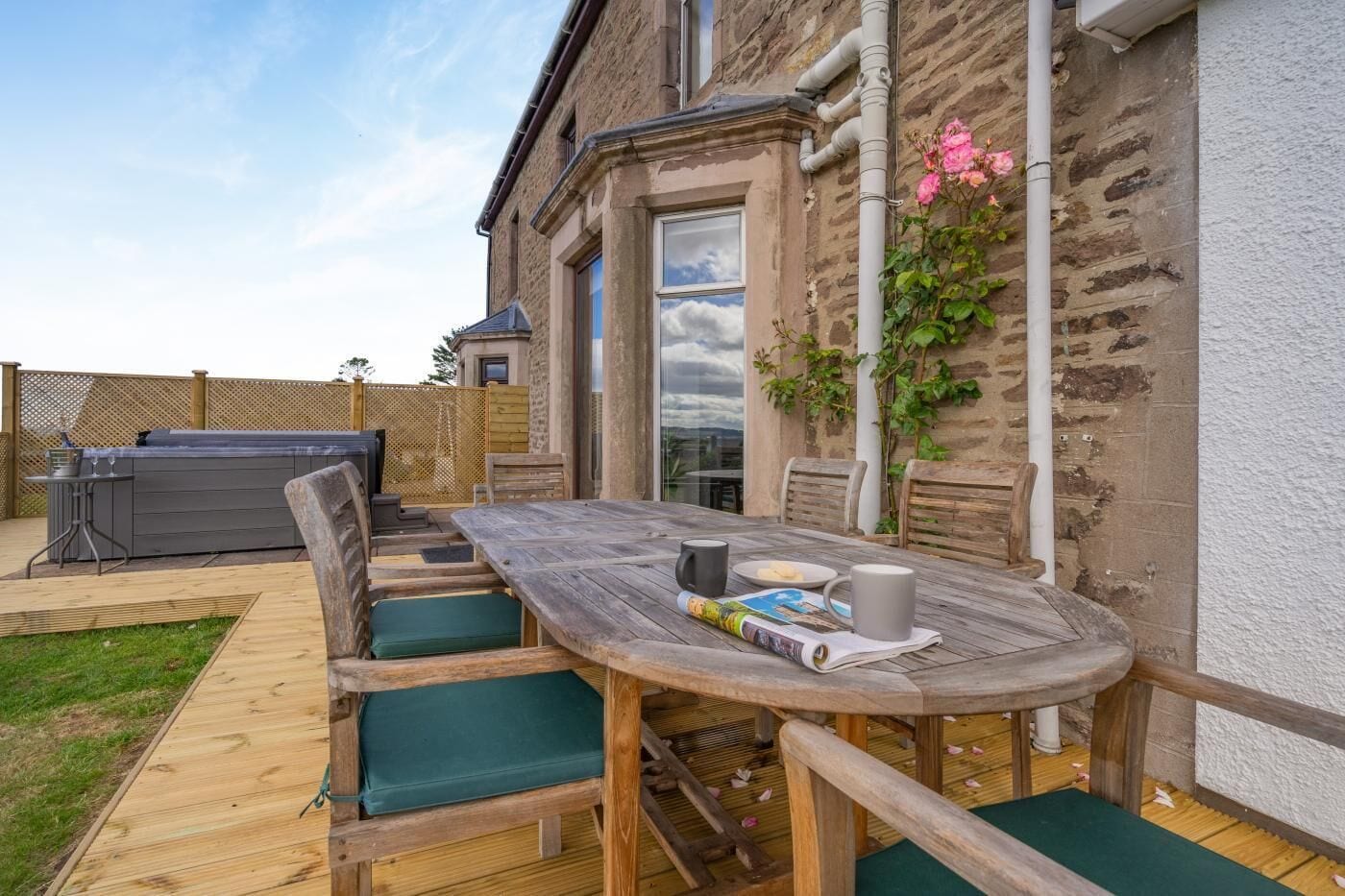 An outdoor patio with a wooden table, green cushioned chairs, and a barbecue grill.