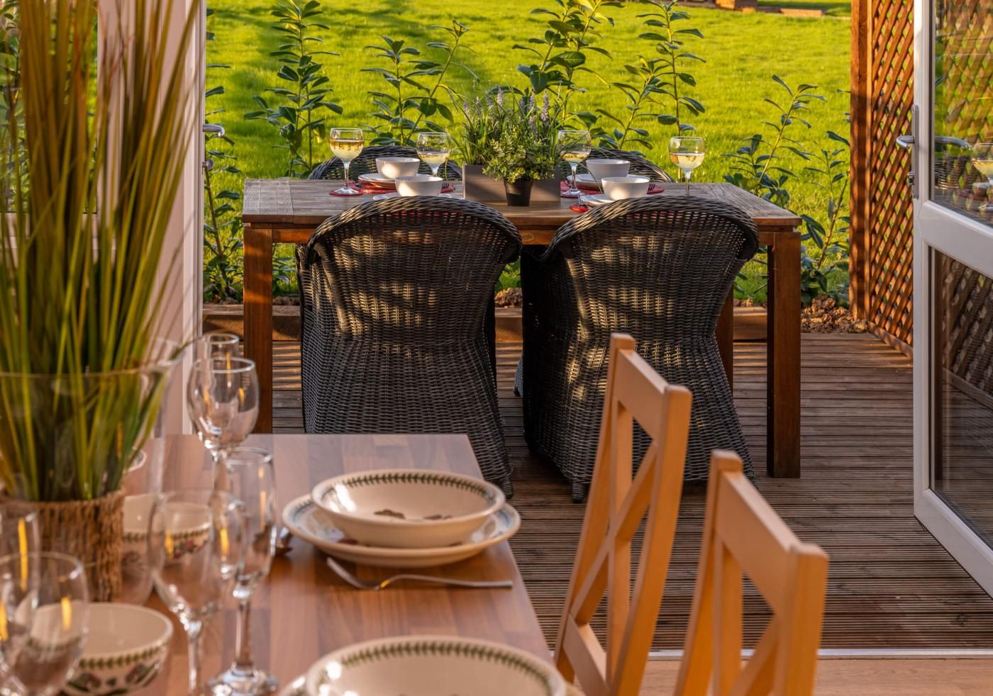 Uma área de refeições ao ar livre com cadeiras de vime e uma mesa de madeira preparada para dois, com vista para um gramado bem cuidado.