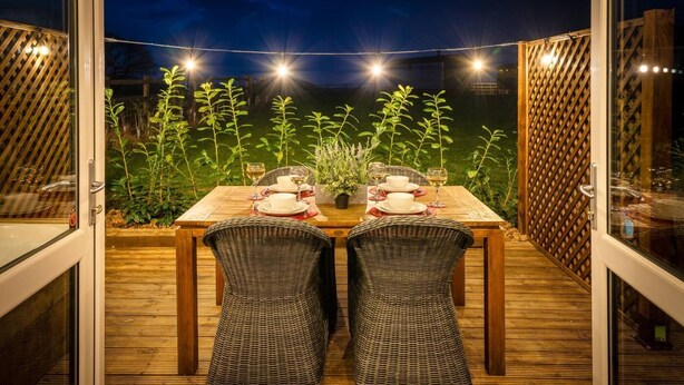 An outdoor dining area with a wooden table and wicker chairs, surrounded by a wooden deck and a view of a garden. - 2 Bed in Wichenford (oc-t45441) (Ockeridge)