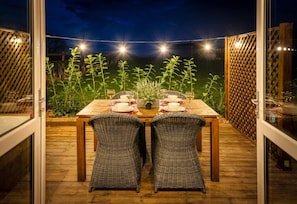 An outdoor dining area with a wooden table and wicker chairs, surrounded by a wooden deck and a view of a garden.