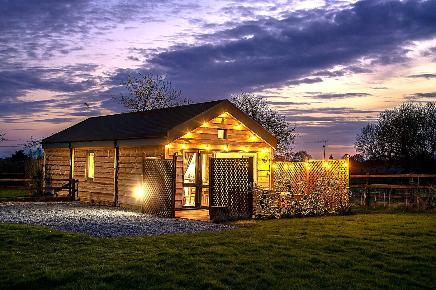 A log cabin with a porch, a fenced area with bicycles, and a gravel driveway.