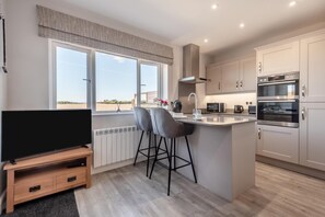 A modern kitchen with a large island, bar stools, a built-in oven, and a television on a wooden stand. - 1 Bed in Thursby (oc-b45540) (Bank End)