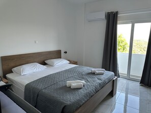 Ein Hotelzimmer mit Bett, Nachttisch, Wand-Klimaanlage und Blick durch ein mit Vorhängen versehenes Fenster.