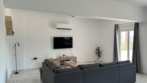 Uma sala de estar moderna com um amplo sofá cinza, uma televisão de tela plana na parede e uma pequena mesa de madeira lateral.