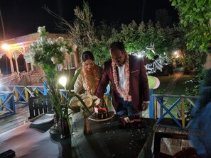 Um casal corta um bolo em um evento ao ar livre, com uma mesa decorada e um jardim ao fundo.