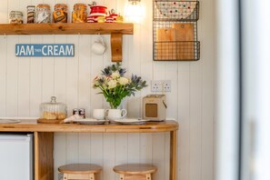 Eine Küche mit einer Holzausziehtheke, einem Schild mit der Aufschrift 'JAM THEN CREAM', Gläsern mit Marmelade, einem Radio und einer Blumenvase.