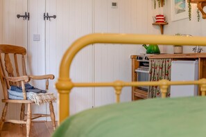 Ein Bettgestell aus Holz, eine grüne Bettdecke, ein Holzsessel mit Decke und Schuhen, ein kleiner Tisch mit einer grünen Vase und ein weißer Kühlschrank.