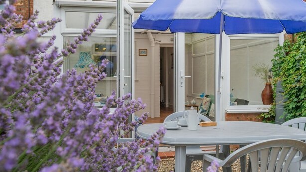 An outdoor seating area with a table and chairs, a blue umbrella, and a view of a house with a glass door. - 2 Bed in Snettisham (oc-d45324) (Snettisham)