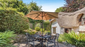 An outdoor patio with a round table and chairs, a large umbrella, and a thatched-roof building. - 3 Bed in Combe St. Nicholas (oc-s45333) (Combe Saint Nicholas)