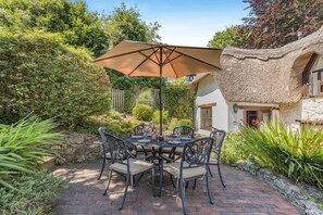 Eine Terrasse im Freien mit einem runden Tisch, Stühlen, einem großen Sonnenschirm und einem Gebäude mit Strohdach.