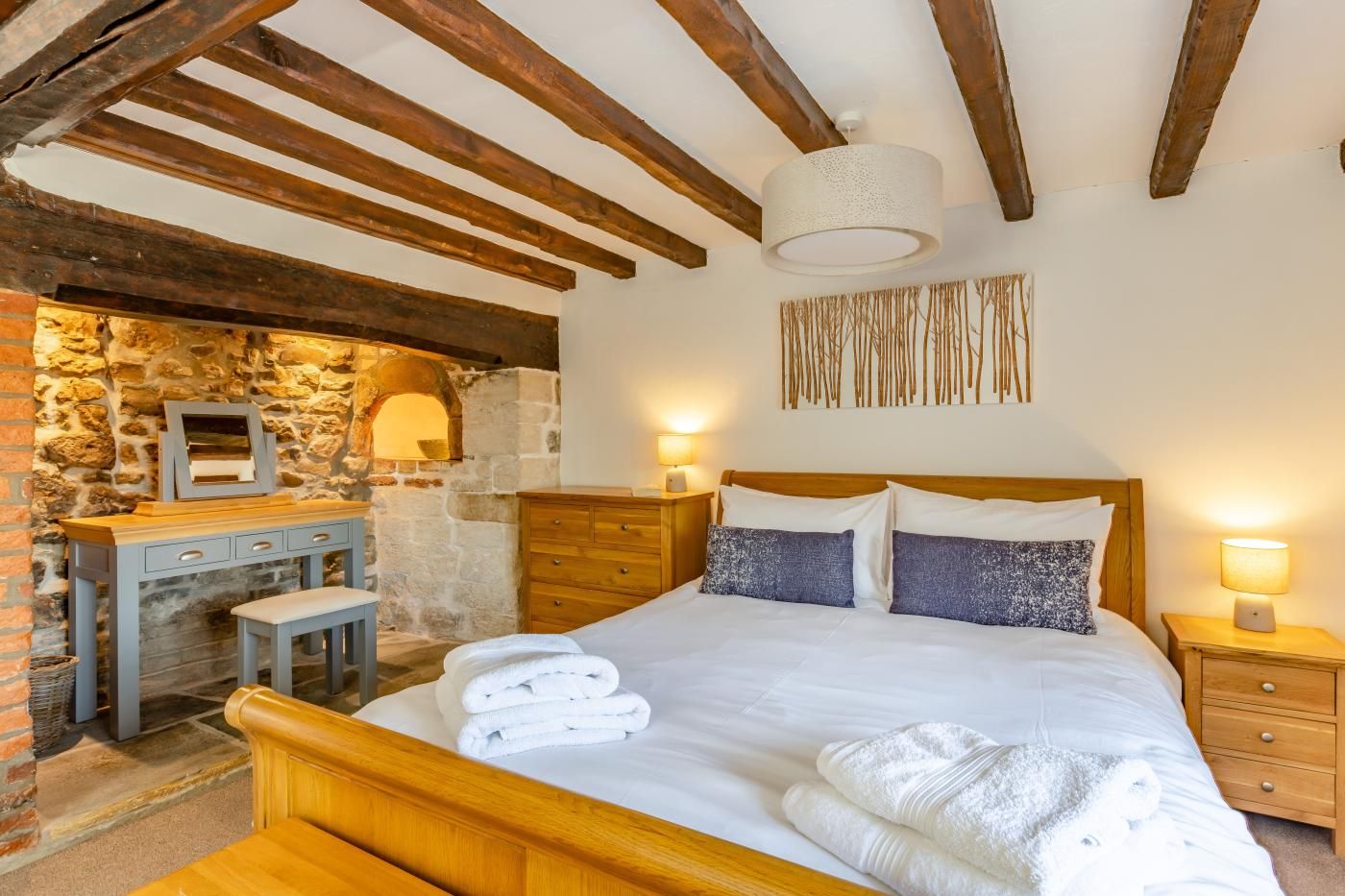 A bedroom with a stone wall, wooden ceiling beams, a bed with white linens, a wooden nightstand, and a desk with a mirror.