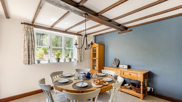 A dining area with a round table set for four, surrounded by white chairs. A wooden cabinet with glass doors is against the wall, and a window with curtains is visible. - 3 Bed in Combe St. Nicholas (oc-s45333) (Combe Saint Nicholas)