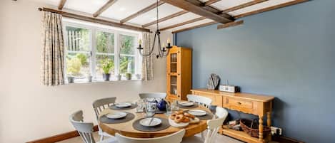 Uma sala de jantar com uma mesa redonda posta para quatro pessoas, cercada por cadeiras brancas. Um armário de madeira com portas de vidro está encostado na parede, e uma janela com cortinas é visível.