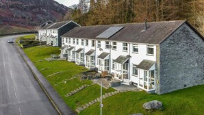 A row of white buildings with stone facades, a paved road, and a grassy area with rocks. - 2 Bed in Ambleside (oc-lr114) (Chapel Stile)