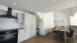 A modern kitchen with a stove, cabinets, and a chimney. There is a dining area with a table set for four and a sofa. - 5 Bed in Llandderfel (oc-b45562) (Llandderfel)