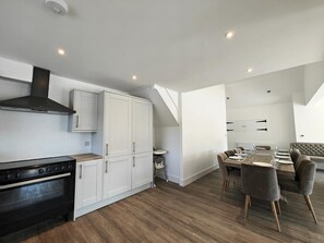 Eine moderne Küche mit Herd, Schränken und einem Schornstein. Es gibt einen Essbereich mit einem Tisch für vier Personen und einer Couch.