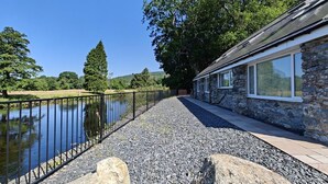 A stone building with a gravel pathway leading to a body of water, surrounded by trees and a metal fence. - 5 Bed in Llandderfel (oc-b45562) (Llandderfel)