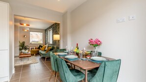 Un comedor con una mesa de madera preparada para cuatro personas, sillas tapizadas de verde y un florero con flores rosadas.