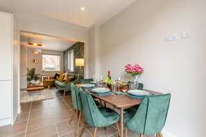 Ein Essbereich mit einem Holztisch, gedeckter Platz für vier Personen, grünen Polsterstühlen und einer Vase mit rosa Blumen.