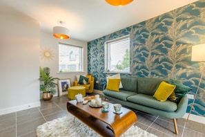 Ein Wohnzimmer mit einem grünen Sofa, gelben Kissen, einem Holztisch und einer Wand mit Muster.