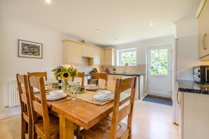 A dining area with a wooden table set for four, surrounded by chairs, with a vase of sunflowers on the table. - 4 Bed in Newquay (oc-s45403) (Black Cross)