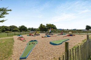 A playground with various play structures, including slides and climbing areas, surrounded by a fence and trees. - 4 Bed in Newquay (oc-s45403) (Black Cross)