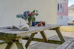 A wooden picnic table with an open book, a vase with flowers, and a cup with strawberries. - 4 Bed in Newquay (oc-s45403) (Black Cross)