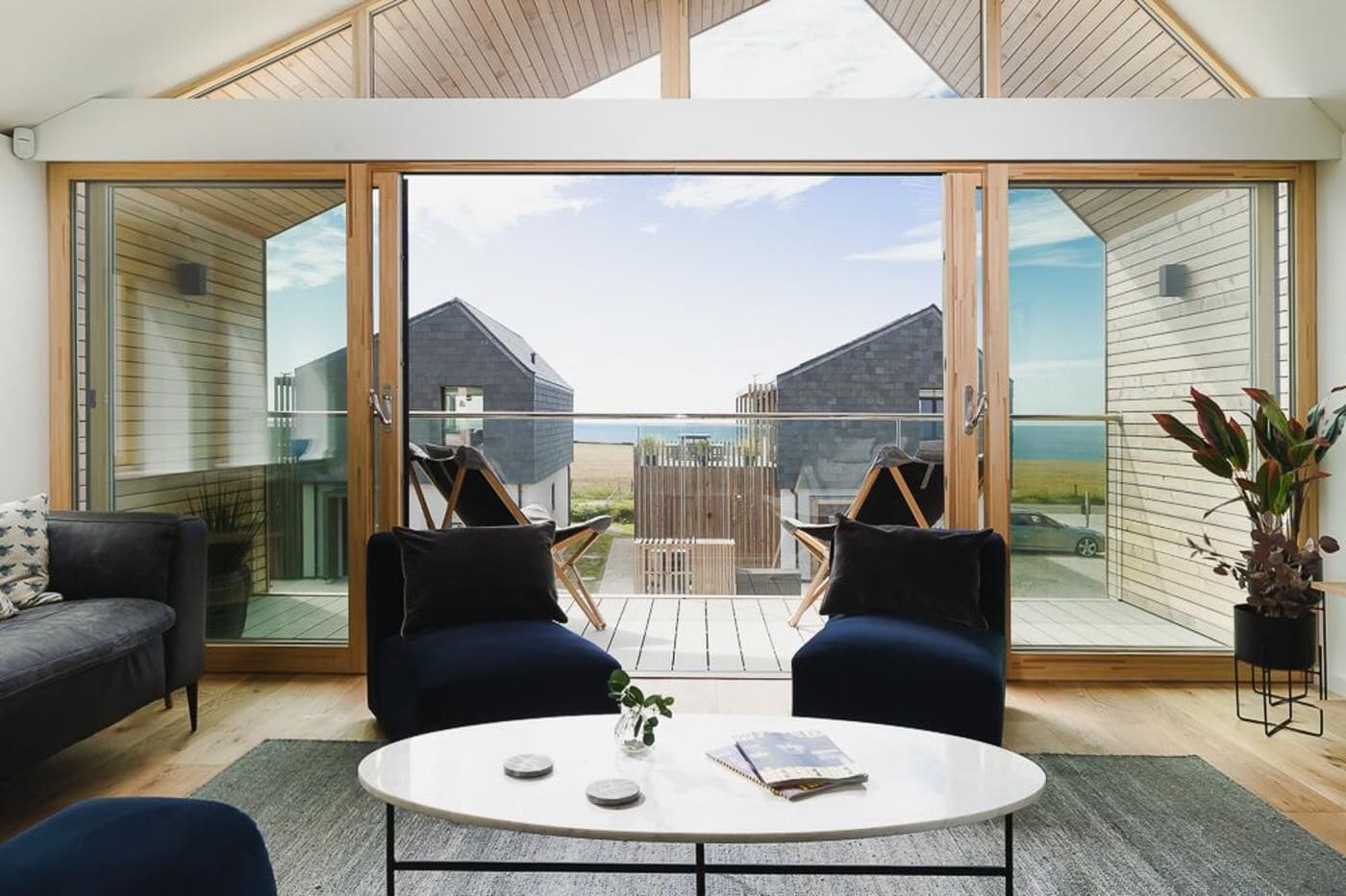 A modern living room with a glass wall, two dark sofas, a round coffee table, and a view of a beach and buildings.
