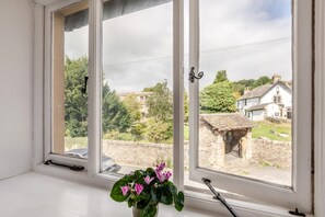 Ein Fenster mit Blick auf eine Steinmauer und traditionelle Gebäude, eine Topfpflanze mit rosa Blüten und ein Metallgriff.