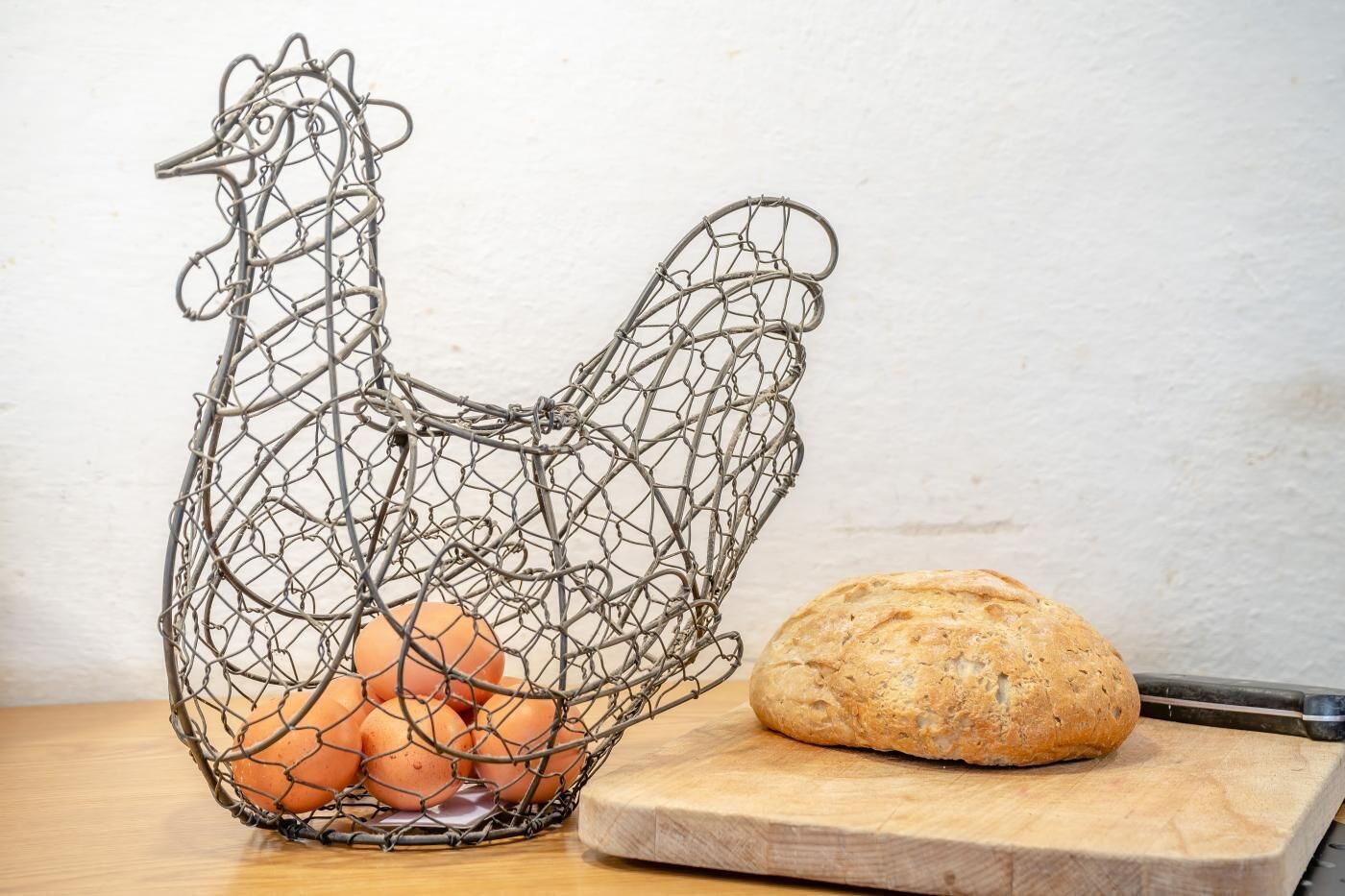 Ein korb in Form eines Hühnchens mit Eiern, einem Brotlaib und einem Schneidebrett auf einer Holzoberfläche.