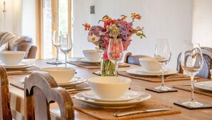 Ein Esstisch ist gedeckt mit weißen Schüsseln, Tellern und Weingläsern, eine Vase mit Blumen und ein hölzerner Stuhl.